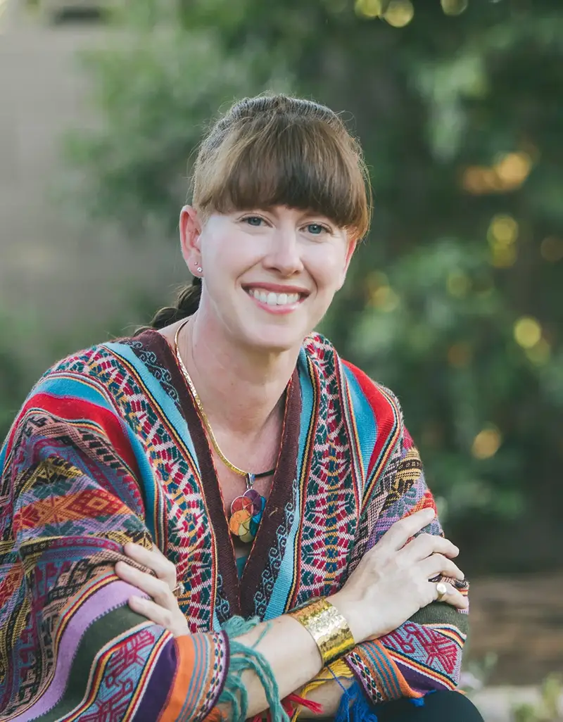 Portrait of a smiling young woman with brown hair wearing a colorful geometric patterned jacket and layered bracelets, photographed outdoors.
