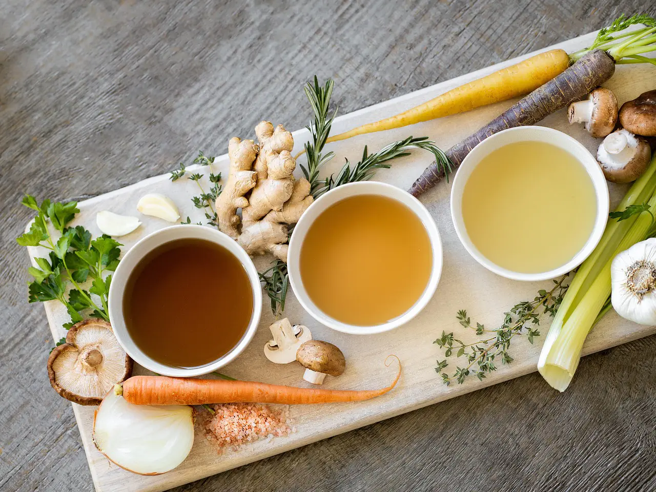 Three white bowls containing different colored broths on a wooden board surrounded by fresh vegetables, herbs, ginger, and salt. Image by Bluebird Provisions from Pixabay