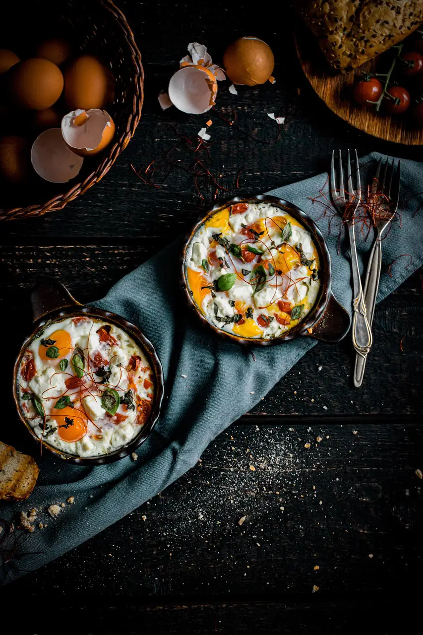 Two cast iron skillets with baked eggs, white sauce, and fresh herbs, surrounded by eggshells, forks, and baskets of eggs and tomatoes. Image by Renata Tlałka from Pixabay
